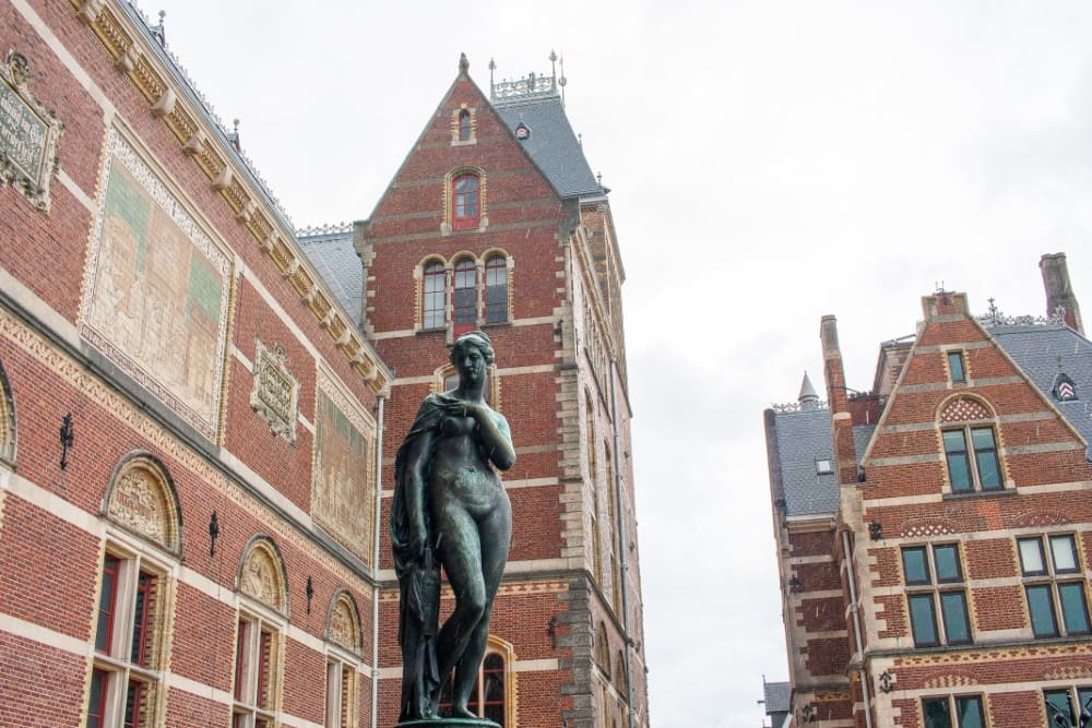A bronze statue of a woman stands in front of the Rijksmuseum, with red-brick architecture and cloudy skies in the background.
