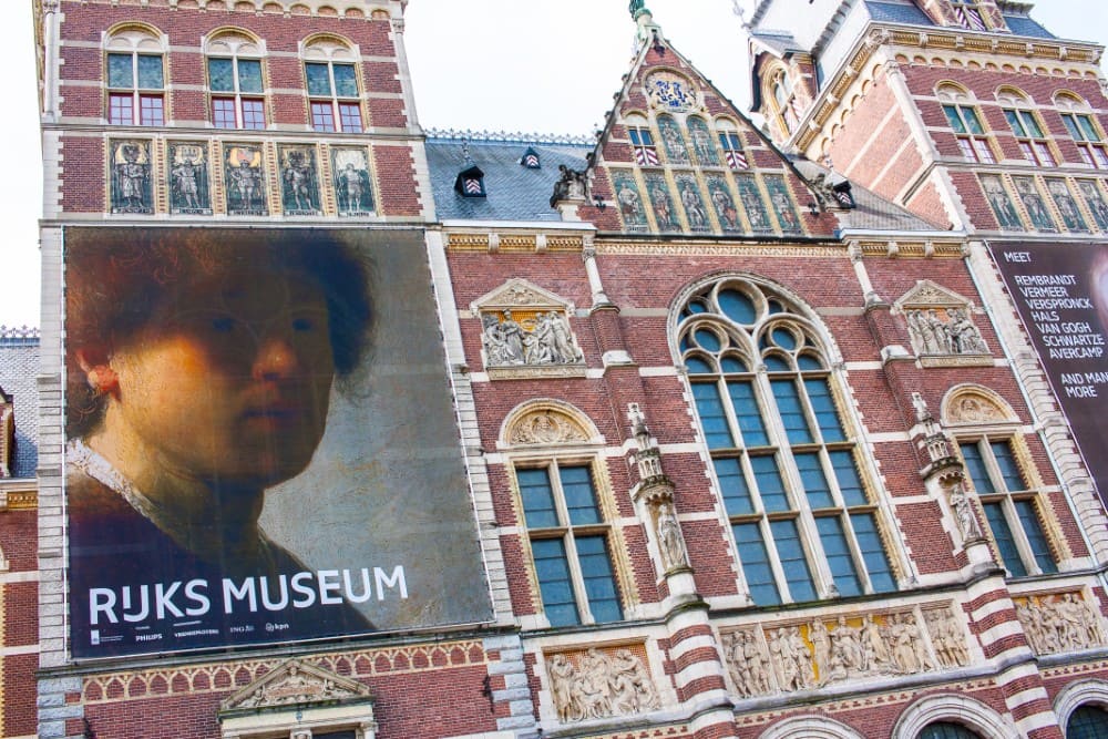 Exterior view of the Rijksmuseum in Amsterdam, featuring a large banner of a painting by Rembrandt on the façade, with intricate architectural details and decorative elements.