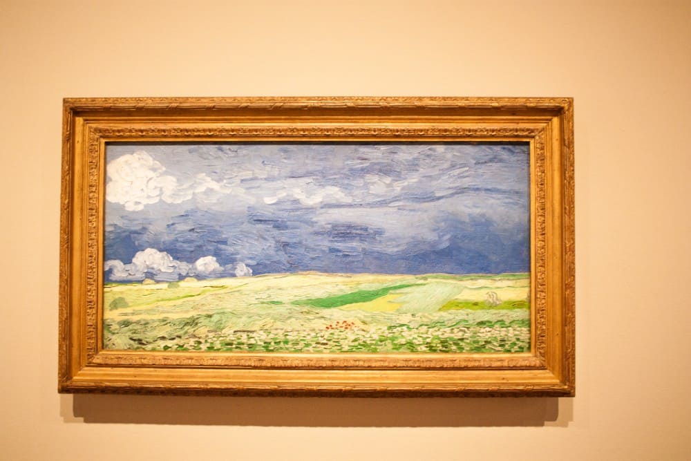At the Van Gogh Museum in Amsterdam: A framed painting depicting a landscape with blue skies, white clouds, and green fields.